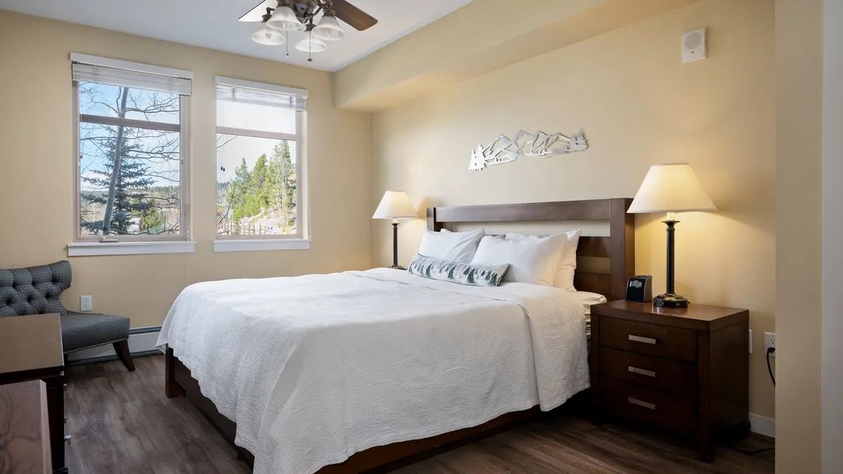 A peaceful bedroom scene featuring a well-made bed with plush bedding, a nightstand, two chairs, and windows letting in natural light. The overall mood is serene and restful, ideal for unwinding after an active day.
