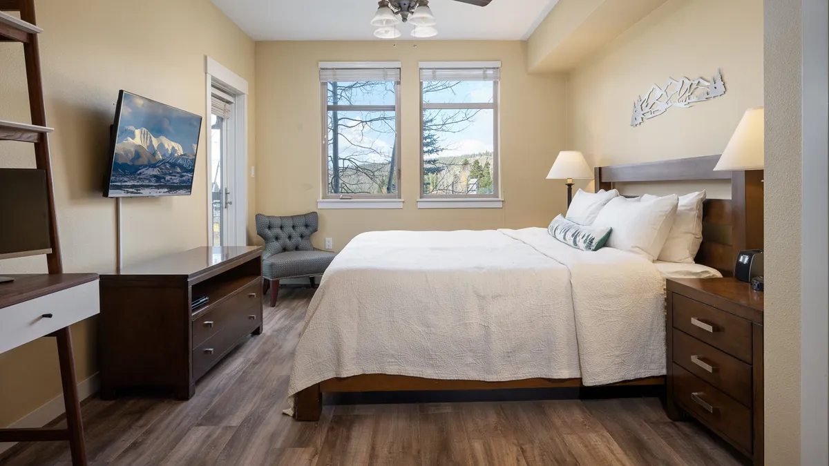 A comfortable bedroom with a bed, a television, a chair by the window, and tasteful decor. The window offers a view, and the room feels bright and inviting, ideal for relaxing, watching TV, or enjoying a quiet morning.
