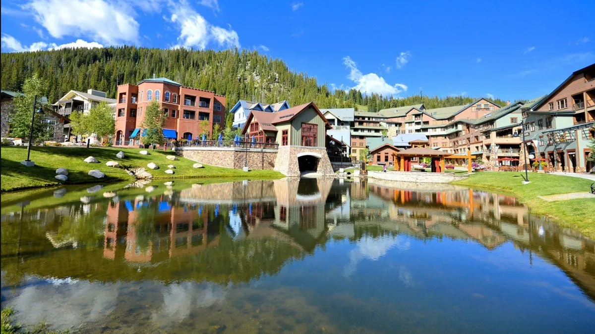 This vibrant exterior scene shows a pond bordered by a pedestrian bridge, colorful buildings, and leafy trees. People can be seen strolling along the water, picnicking on the grass, and children are playing in the playground, evoking a lively, family-friendly resort atmosphere in the summer.