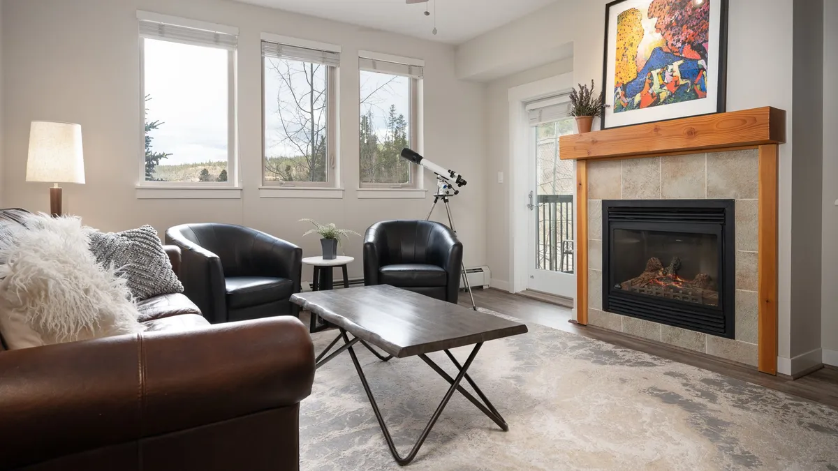 The image showcases a warm, inviting living space with a glowing fireplace, plush sofa, tasteful artwork, and large windows framing serene mountain views. Guests are seen relaxing, socializing, and enjoying cozy moments indoors while nature beckons just beyond the glass.