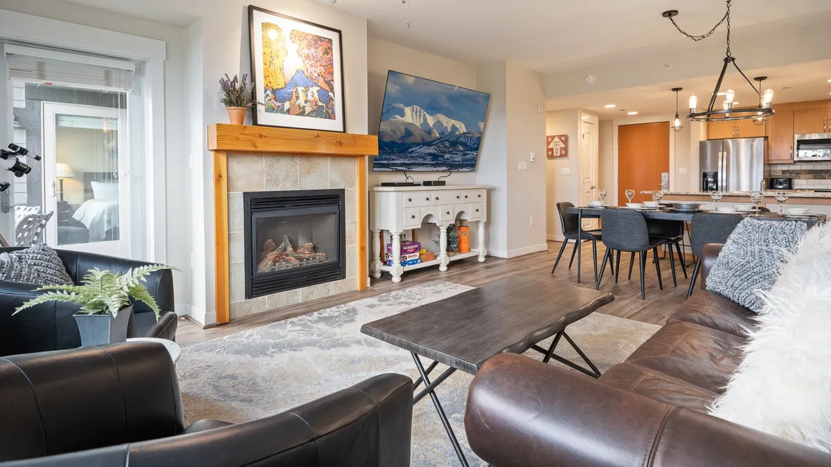 A warm, inviting great room featuring a stone gas fireplace, a mounted television, a contemporary dining table, and tasteful wall art. The room is spacious with comfortable seating and an open layout that fosters connection and relaxation.