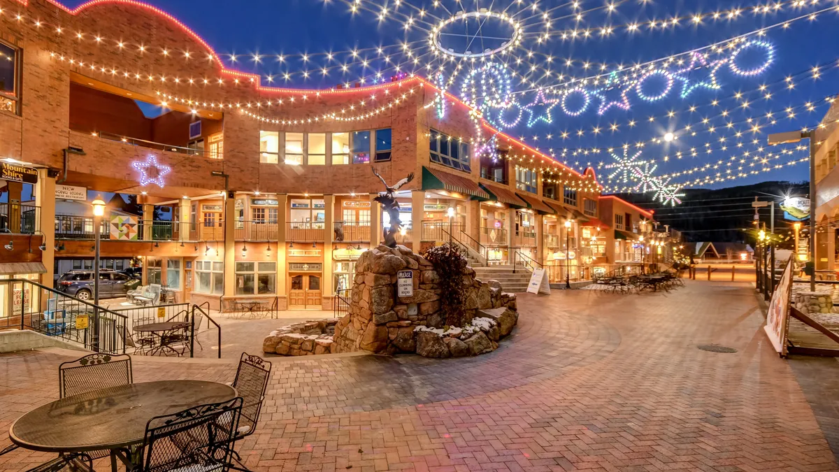 A bustling exterior scene of downtown Winter Park, with decorative lights, a stone fountain, outdoor seating, and shopfronts. The atmosphere is festive, inviting visitors to enjoy dining, shopping, and evening entertainment.