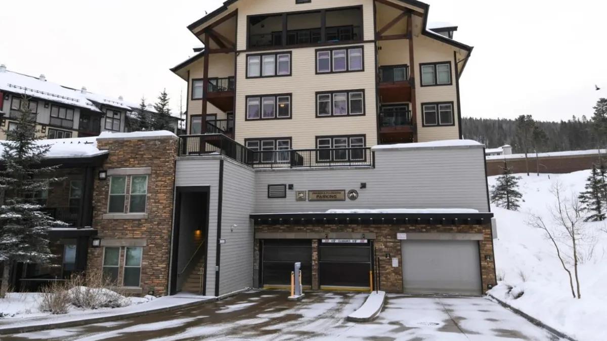 An exterior winter view of a multi-story lodge-style building with a stone façade, surrounded by snow and mountains. The scene suggests a warm, welcoming atmosphere with the buzz of guests returning from a day on the slopes.