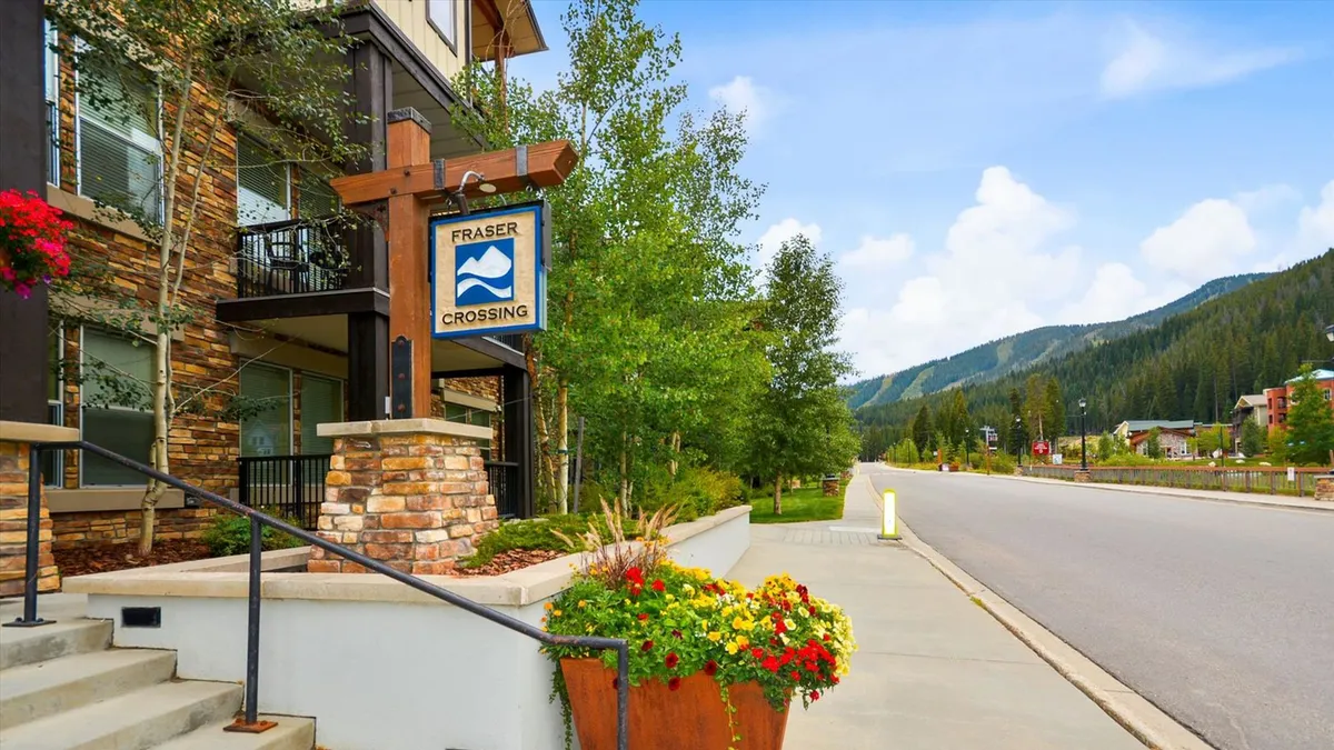 The image showcases the Fraser Crossing sign with stone architecture, flower planters, and surrounding greenery. The outdoor setting feels welcoming and hints at the connection to local trails and nature, with paths leading into the lush landscape.