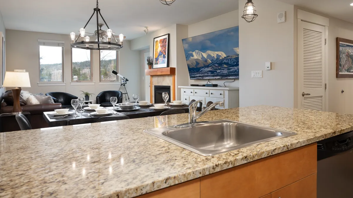 An open-plan space combining a modern kitchen, dining table, fireplace, and television. The kitchen sink overlooks the dining and living area, and mountain-inspired art complements the space, reinforcing the alpine theme.