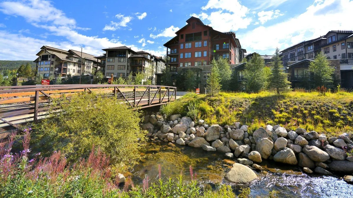 This exterior image features a picturesque wooden bridge crossing a flowing stream, surrounded by landscaped greenery and modern buildings in the background. People can be seen walking and relaxing by the water, with vibrant foliage showcasing the seasonal beauty of the area.