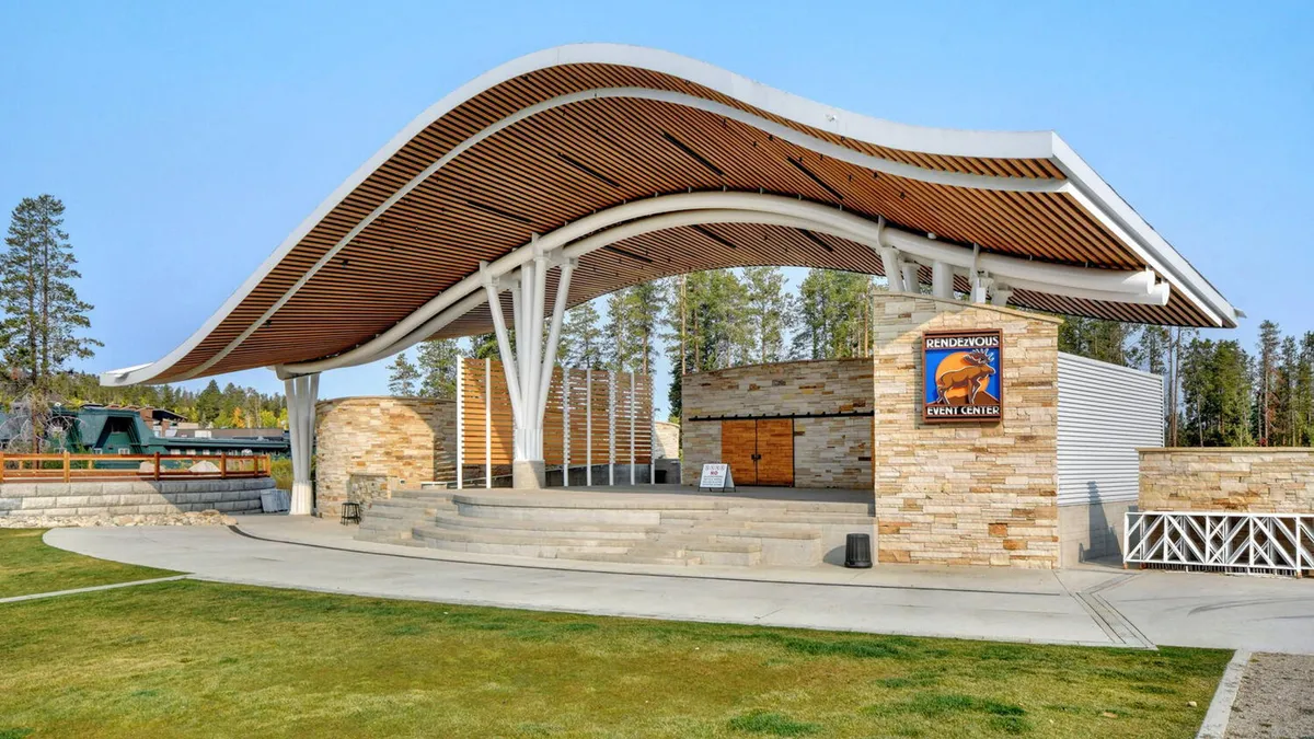 An exterior shot of the local event center, featuring a curved roof structure, stone façade, and a visible event center sign. The space is designed for community gatherings, outdoor performances, and festive local events.